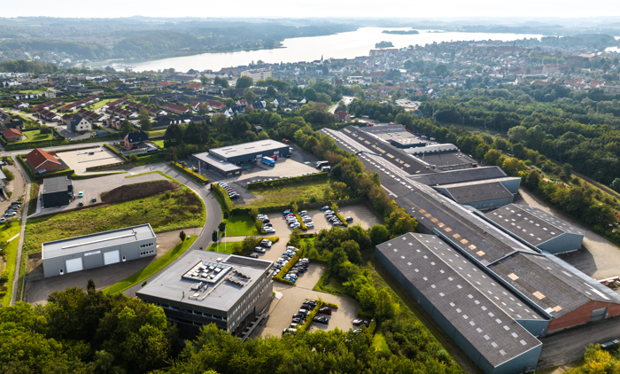 Luftfoto af et byområde med en kombination af boligkvarterer, erhvervsbygninger og grønne områder. I forgrunden ses flere industribygninger og parkeringspladser omgivet af grønne træer, mens der i baggrunden er huse og en sø, som strækker sig i landskabet. Billedet giver et overblik over både det beboede og kommercielle område, der grænser op til naturlige omgivelser