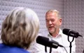 Mand med gråt skæg i hvid skjorte taler i podcaststudie med mikrofoner; medvært set bagfra, akustikpaneler i baggrunden.